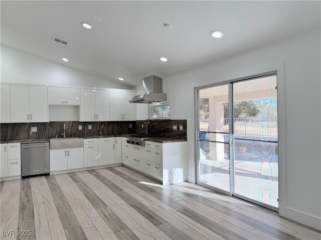 kitchen featuring lofted ceiling, wall chimney range hood, sink, dishwasher, and white cabinets