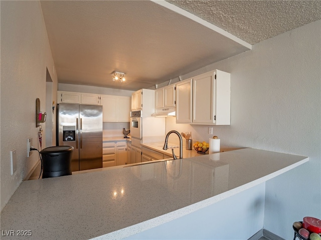 kitchen with wall oven, a textured wall, a peninsula, under cabinet range hood, and stainless steel refrigerator with ice dispenser