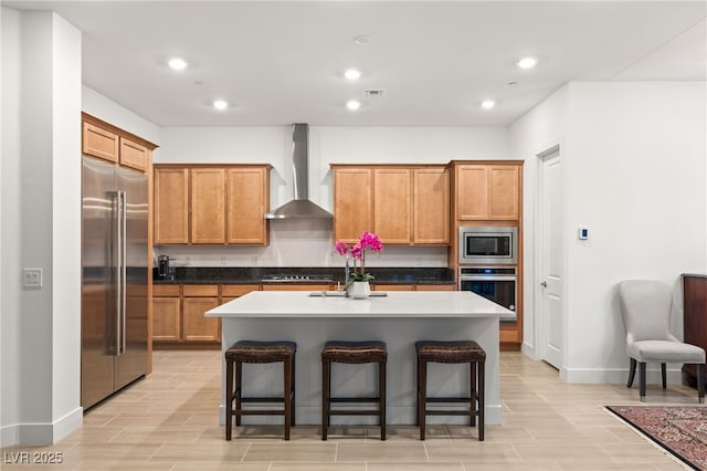 kitchen featuring built in appliances, wall chimney range hood, an island with sink, and a kitchen bar