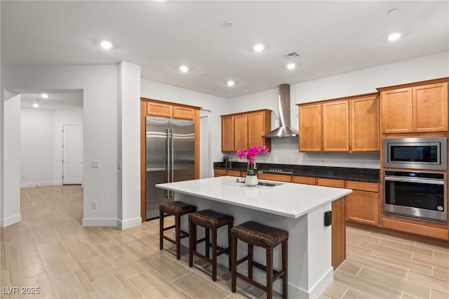 kitchen with an island with sink, built in appliances, a kitchen breakfast bar, and wall chimney exhaust hood