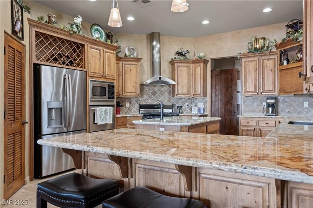 kitchen with pendant lighting, wall chimney exhaust hood, light stone countertops, appliances with stainless steel finishes, and a kitchen bar