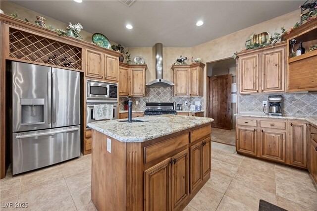 kitchen with a center island with sink, wall chimney range hood, tasteful backsplash, light stone counters, and stainless steel appliances