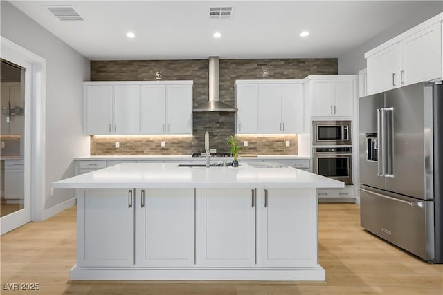 kitchen featuring white cabinetry, an island with sink, appliances with stainless steel finishes, and wall chimney exhaust hood