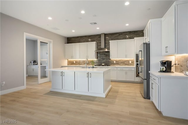 kitchen featuring light wood-type flooring, stainless steel refrigerator, wall chimney range hood, a kitchen island with sink, and white cabinets