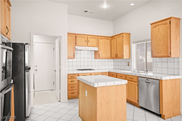 kitchen with tile counters, appliances with stainless steel finishes, light tile patterned flooring, a kitchen island, and backsplash