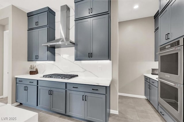 kitchen featuring light stone countertops, wall chimney exhaust hood, light tile patterned floors, backsplash, and appliances with stainless steel finishes