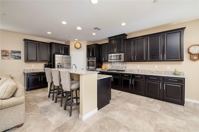 kitchen with appliances with stainless steel finishes, an island with sink, a breakfast bar, light stone counters, and tasteful backsplash