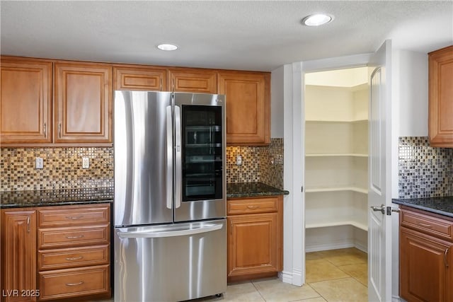 kitchen with stainless steel refrigerator, dark stone countertops, light tile patterned floors, and decorative backsplash