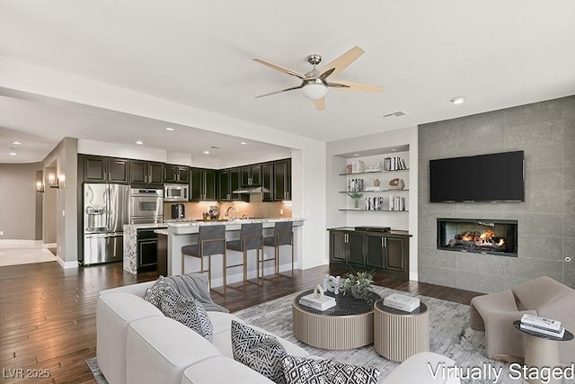 living room featuring sink, a tiled fireplace, ceiling fan, built in features, and dark hardwood / wood-style flooring