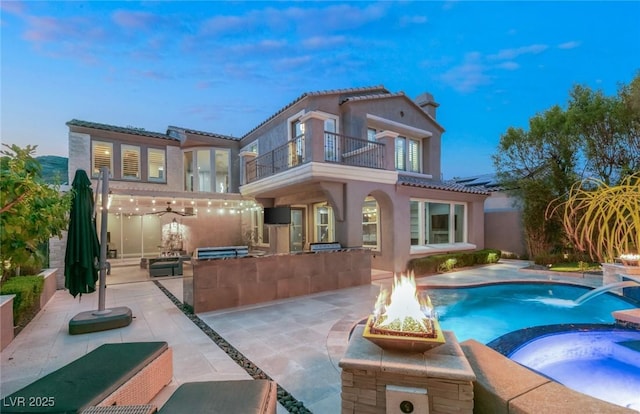 back house at dusk with an outdoor fire pit, a balcony, an in ground hot tub, pool water feature, and a patio area