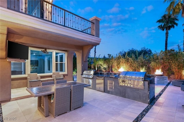 patio terrace at dusk featuring an outdoor kitchen, a balcony, and a grill