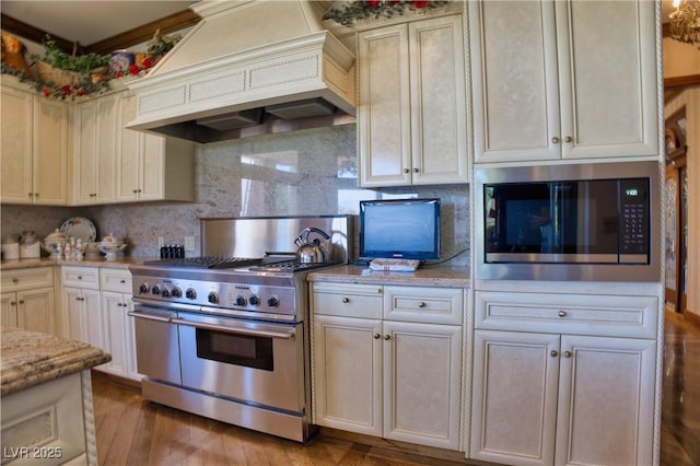 kitchen with light stone counters, custom exhaust hood, backsplash, appliances with stainless steel finishes, and cream cabinetry