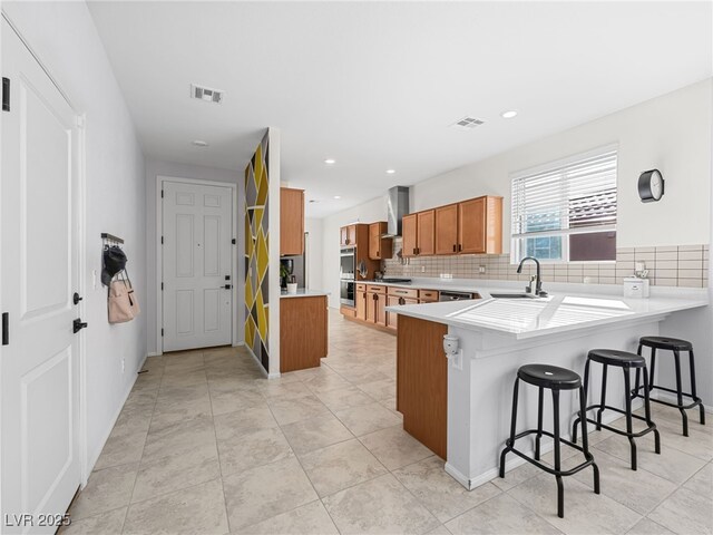 kitchen featuring sink, a kitchen bar, tasteful backsplash, kitchen peninsula, and wall chimney range hood