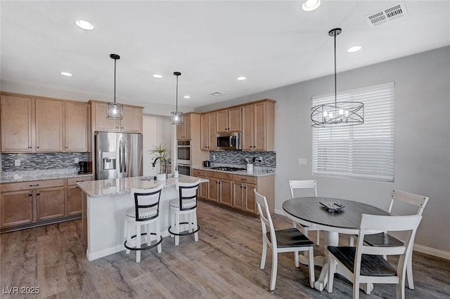 kitchen with stainless steel appliances, light stone counters, hardwood / wood-style flooring, an island with sink, and hanging light fixtures