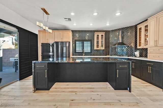 kitchen featuring a kitchen island with sink, black appliances, hanging light fixtures, light hardwood / wood-style floors, and wall chimney exhaust hood