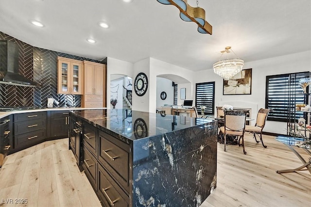 kitchen featuring light wood-type flooring, glass insert cabinets, arched walkways, wall chimney exhaust hood, and cooktop