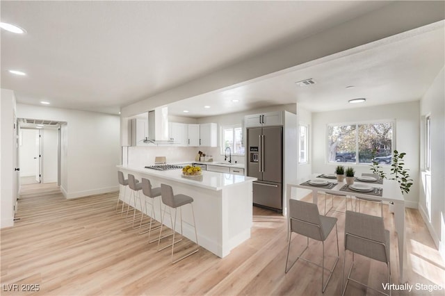 kitchen featuring white cabinets, wall chimney exhaust hood, high quality fridge, light wood-type flooring, and a breakfast bar