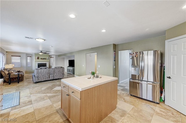 kitchen featuring a tiled fireplace, a center island, ceiling fan, stainless steel fridge, and light brown cabinetry