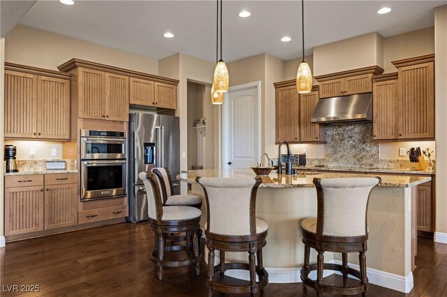 kitchen featuring stainless steel appliances, a center island with sink, hanging light fixtures, and light stone countertops