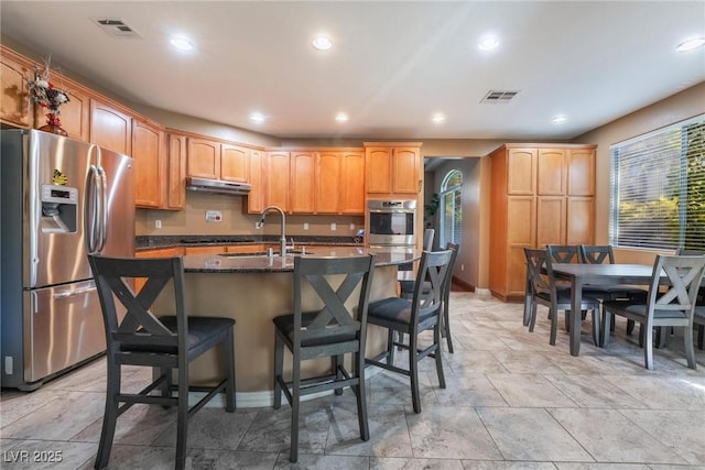 kitchen featuring stainless steel appliances, sink, a kitchen bar, an island with sink, and dark stone counters