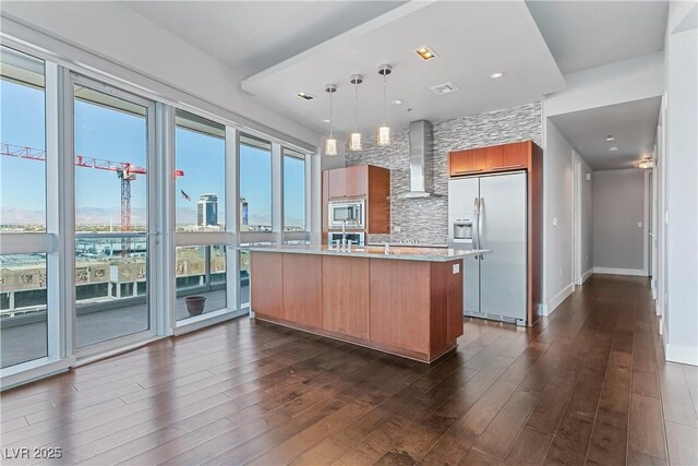 kitchen with built in appliances, wall chimney exhaust hood, hanging light fixtures, a kitchen island with sink, and dark wood-type flooring