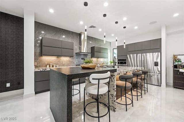 kitchen featuring wall chimney exhaust hood, hanging light fixtures, a kitchen island with sink, a breakfast bar area, and appliances with stainless steel finishes