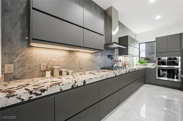 kitchen featuring gray cabinets, appliances with stainless steel finishes, light stone counters, wall chimney exhaust hood, and backsplash