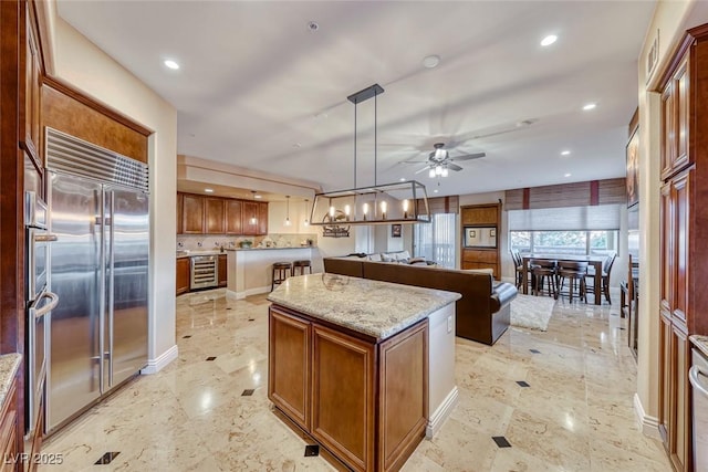 kitchen with hanging light fixtures, built in refrigerator, ceiling fan, light stone counters, and a kitchen island