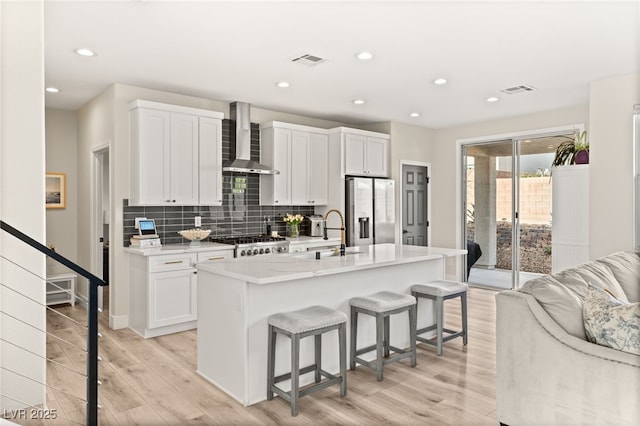 kitchen featuring a center island with sink, wall chimney range hood, a breakfast bar, stainless steel fridge, and white cabinetry