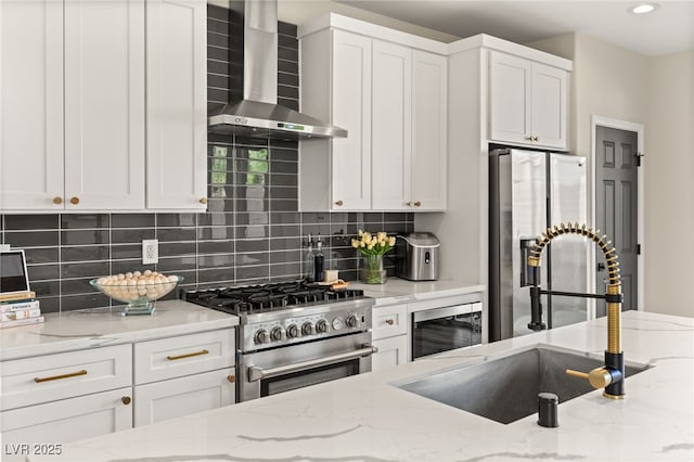 kitchen featuring light stone counters, wall chimney exhaust hood, white cabinetry, appliances with stainless steel finishes, and sink