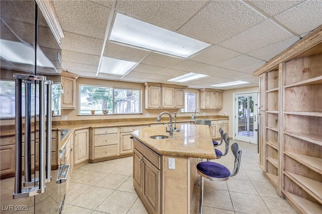 kitchen featuring a kitchen breakfast bar, an island with sink, a drop ceiling, light brown cabinets, and sink