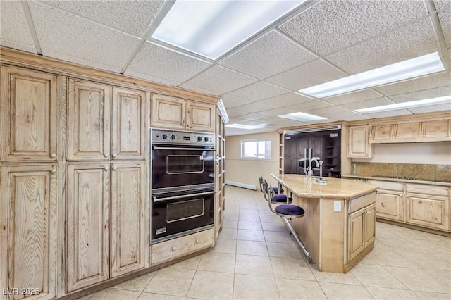 kitchen featuring black built in fridge, double oven, light brown cabinets, and an island with sink