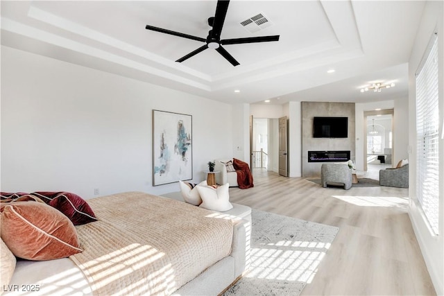 bedroom featuring light hardwood / wood-style floors, a premium fireplace, ceiling fan, and a tray ceiling