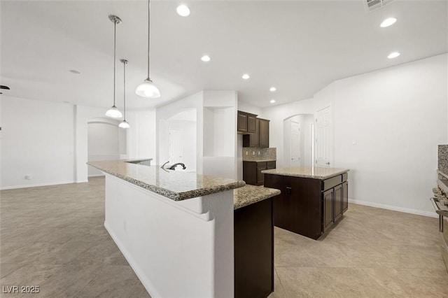 kitchen with light stone countertops, a kitchen island with sink, dark brown cabinets, and hanging light fixtures