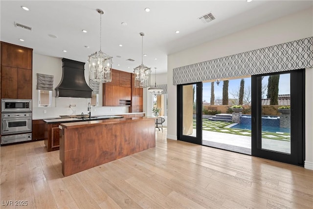 kitchen featuring stainless steel appliances, visible vents, light wood finished floors, a large island with sink, and custom range hood