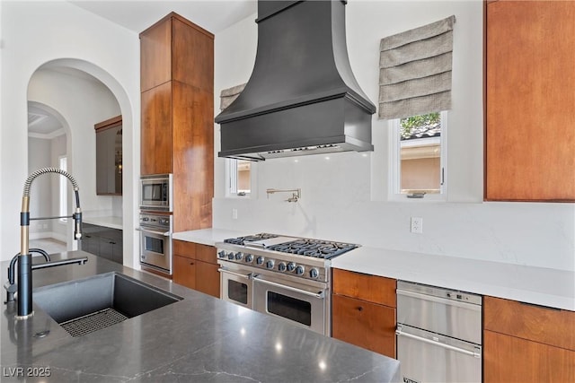 kitchen featuring brown cabinetry, modern cabinets, custom range hood, appliances with stainless steel finishes, and a sink