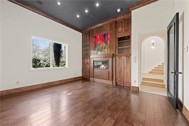 unfurnished living room featuring arched walkways, ornamental molding, dark wood-type flooring, and a glass covered fireplace