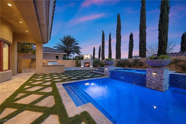 pool at dusk with exterior kitchen, a patio area, fence, and a lit fireplace