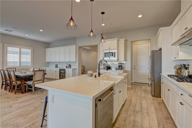 kitchen with hanging light fixtures, a center island with sink, light wood-type flooring, white cabinetry, and appliances with stainless steel finishes