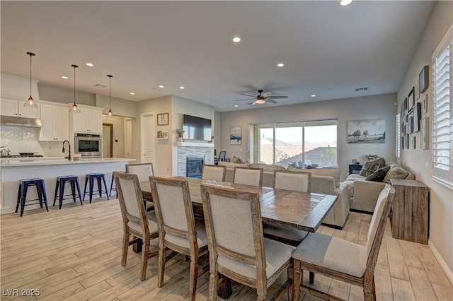 dining room with sink, a stone fireplace, ceiling fan, and light hardwood / wood-style flooring