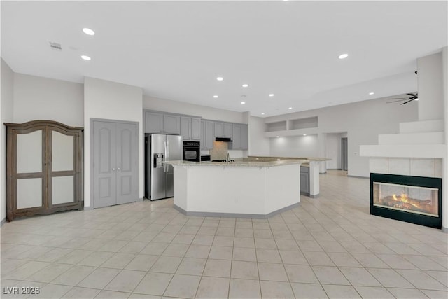 kitchen with gray cabinets, a center island with sink, light stone countertops, black oven, and stainless steel refrigerator with ice dispenser