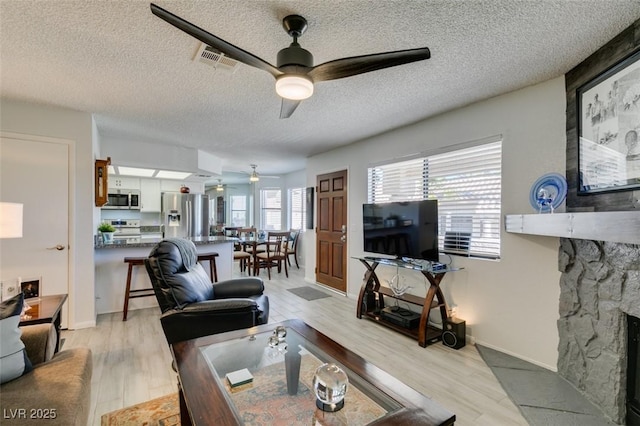 living room with a stone fireplace, a textured ceiling, ceiling fan, and light hardwood / wood-style flooring