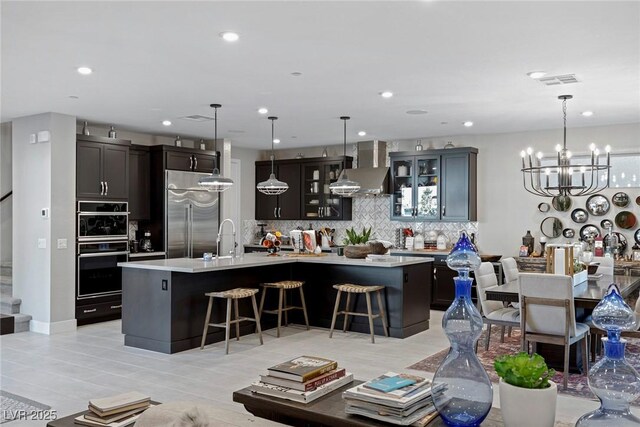 kitchen featuring hanging light fixtures, built in refrigerator, wall chimney range hood, and a center island with sink