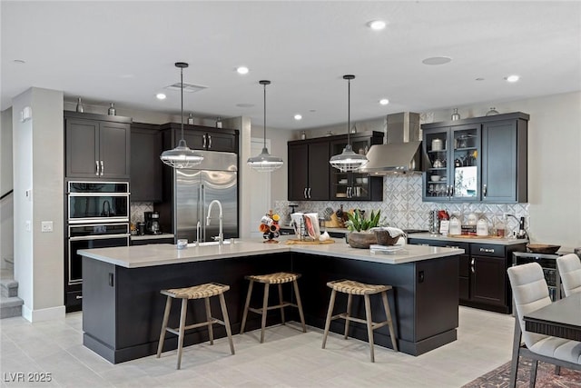 kitchen with built in refrigerator, wall chimney range hood, a kitchen island with sink, and hanging light fixtures