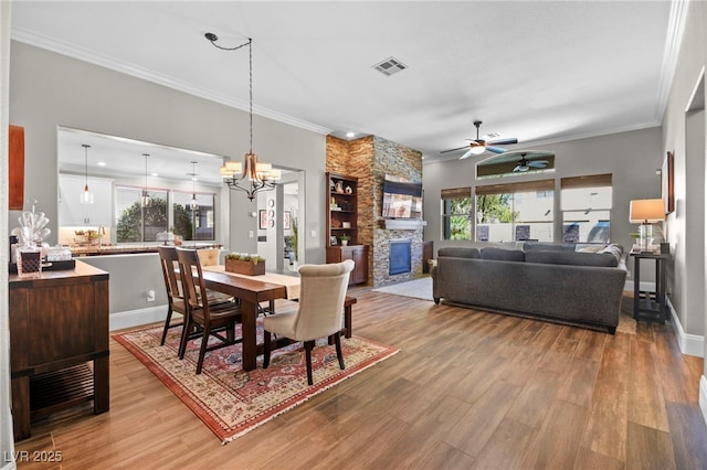 dining area featuring ceiling fan with notable chandelier, light wood-type flooring, crown molding, and a stone fireplace