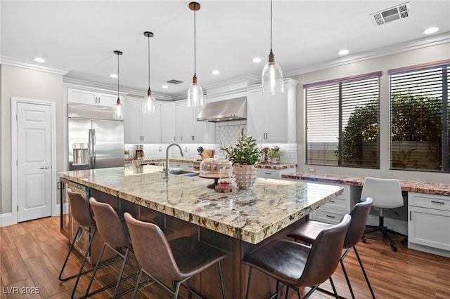 kitchen with hanging light fixtures, an island with sink, sink, white cabinetry, and exhaust hood