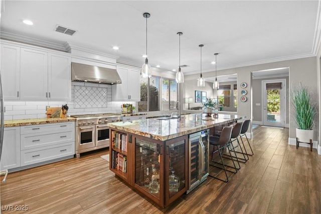 kitchen featuring a center island with sink, wall chimney range hood, range with two ovens, and white cabinetry