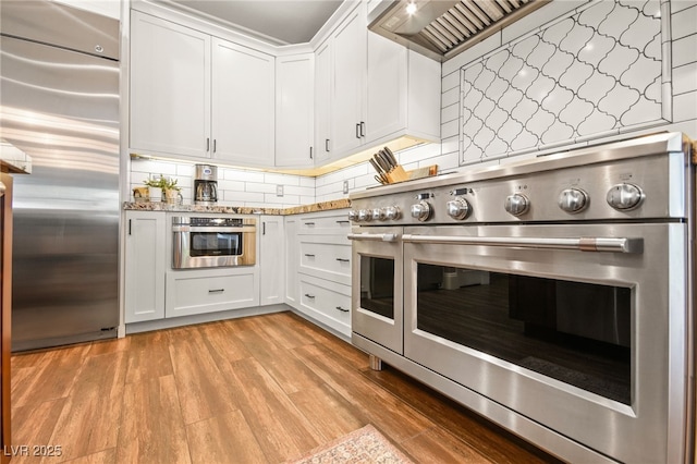 kitchen featuring wall chimney range hood, light hardwood / wood-style floors, decorative backsplash, white cabinetry, and appliances with stainless steel finishes