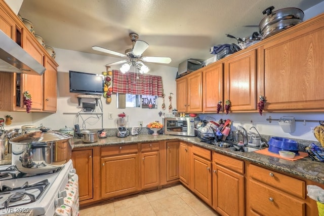 kitchen featuring wall chimney exhaust hood, dark stone counters, light tile patterned flooring, ceiling fan, and sink