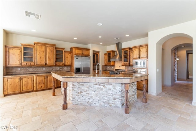 kitchen with a breakfast bar area, a center island with sink, built in appliances, decorative backsplash, and wall chimney range hood